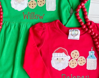 May include: Two Christmas-themed long-sleeved shirts for children. The green shirt has a Santa Claus, milk bottle, and cookies design with the name "Willow" embroidered on it. The red shirt has a similar design with the name "Delaney" embroidered on it.