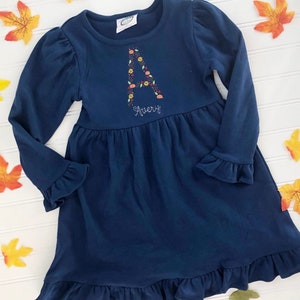 Puede incluir: Un vestido azul marino de manga larga con un dobladillo y puños con volantes. El vestido tiene una letra "A" bordada con flores y el nombre "Avery" bordado debajo.