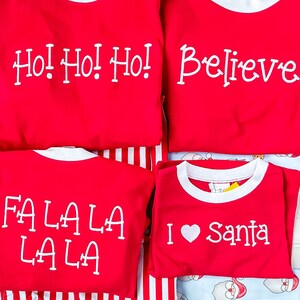 May include: Four red Christmas shirts with white text. The shirts read "Ho! Ho! Ho!", "Believe", "Fa La La La La", and "I &hearts; Santa". The shirts have white collars and are on a striped and Santa-themed background.