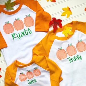Puede incluir: Tres camisetas de béisbol blancas con mangas naranjas presentan calabazas bordadas y nombres. Las camisetas son Ryatt, Teddy y Jack.