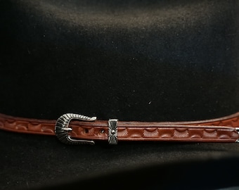 May include: A brown leather hat band with a silver buckle and decorative accents. The band is attached to a black felt hat.