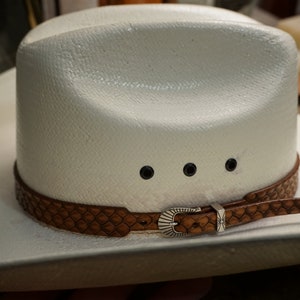 May include: A white cowboy hat with a brown leather band featuring a decorative buckle. The hat has a classic design with a wide brim and a textured surface. Three black eyelets are visible on the crown.