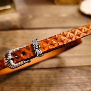 May include: A brown leather belt with a decorative floral pattern and a silver buckle. The belt has a detailed embossed design and a silver tip. The buckle and tip have a vintage, Western-inspired design.