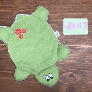 Puede incluir: Un artículo de tela verde con forma de tortuga con una superficie texturizada, detalles de flores rojas y ojos de dibujos animados. Se adjunta una pequeña etiqueta. También hay una tarjeta verde claro con una ilustración de tortuga rosa y el número 7.