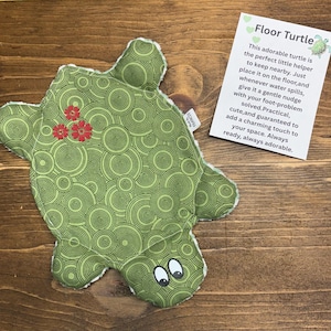 Op de afbeelding: Een schildpadvormige stoffen pad met een groen cirkelpatroon, rode bloemenborduursels en witte ogen. Daarnaast ligt een witte kaart met de tekst: Floor Turtle. This adorable turtle is the perfect little helper to keep nearby. Just place it on the floor, and whenever water spills, give it a gentle nudge with your foot-problem solved. Practical, cute, and guaranteed to add a charming touch to your space. Always ready, always adorable.