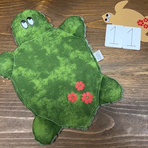 May include: A plush turtle toy featuring a mottled green fabric top with three red flower appliques, white eyes with black pupils, and a small white tag labeled Made by Francis. The turtle has a textured green underside and a fuzzy border.