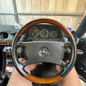 May include: A Mercedes-Benz steering wheel featuring a polished wood rim and black leather grips, centred with a silver three-pointed star emblem. Behind the wheel, the dashboard displays a speedometer, tachometer, and fuel gauge.