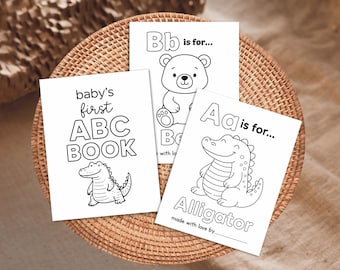 May include: Three white baby ABC books with black outlines are displayed on a woven surface. The books feature illustrations of a bear and an alligator, with the text "baby's first ABC BOOK". The book also says "Bb is for..." and "Aa is for... Alligator".