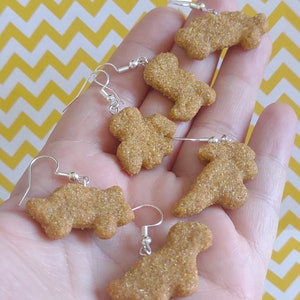 May include: Six novelty earrings in the shape of chicken nuggets. The earrings are a light brown color with a textured surface and silver-colored ear wires. The earrings are displayed against a yellow and white zigzag background.