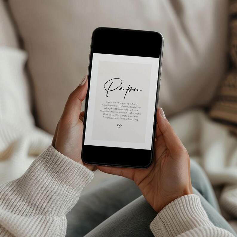 May include: A smartphone displays a digital print with the word "Papa" in elegant script. Below, a list of words is printed in a smaller font. A small heart icon is at the bottom. The phone is held by a person wearing a white sweater.