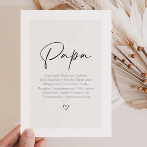 May include: A white card with the word "Papa" in elegant black script. Below, a list of German words describes a father's roles, including "Superhero" and "Motivator." A small heart is at the bottom. The card is held by a hand.