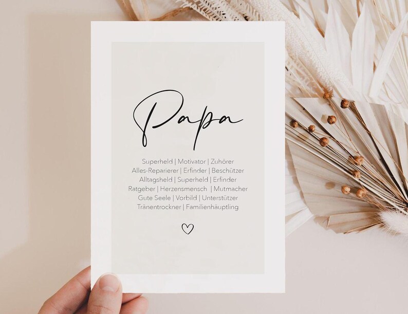 May include: A white card with the word "Papa" in elegant black script. Below, a list of German words describes a father's roles, including "Superhero" and "Motivator." A small heart is at the bottom. The card is held by a hand.