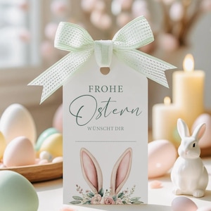 May include: A white tag with the text "FROHE OSTERN" and "WÜNSCHT DIR" in green script, adorned with watercolor bunny ears and flowers. A light green ribbon bow is tied at the top. Easter-themed decor includes eggs and a ceramic bunny.