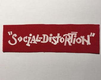 Puede incluir: Un parche rectangular de tela roja con el texto bordado en blanco "SOCIAL-DISTORTION" en una fuente estilizada y dentada. El parche tiene una textura tejida visible y bordes limpios, presentando un contraste marcado entre el fondo rojo y las letras blancas.