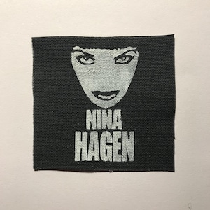 May include: A square black fabric patch featuring a white screen-printed graphic of a face with dark hair and the text NINA HAGEN printed in a bold, sans-serif font below the face. The patch has a visible woven texture throughout the black surface.