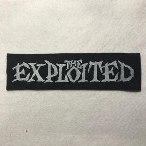 May include: A rectangular black fabric patch featuring the word THE EXPLOITED embroidered in a jagged, light gray, distressed-style font. The lettering is centered on the black background, creating a high-contrast design for this textile accessory.