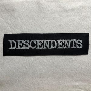 Könnte beinhalten: Ein schwarzer rechteckiger Aufnäher mit dem Wort "DESCENDENTS" in einer verblassten, silberfarbenen Schrift. Der Aufnäher ist wahrscheinlich aus Stoff und zum Aufnähen oder Aufbügeln auf Kleidung oder Accessoires geeignet.