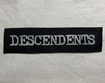 Puede incluir: Un parche rectangular negro con la palabra "DESCENDENTS" en una fuente plateada desgastada. El parche es probablemente de tela y es adecuado para coser o planchar en ropa o accesorios.