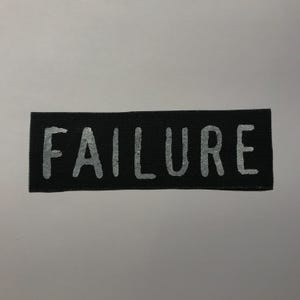 May include: A rectangular black fabric patch featuring the word FAILURE printed in a light gray, distressed, sans-serif font. The patch has a woven texture and clean, straight edges, presenting a high-contrast design on a solid background.