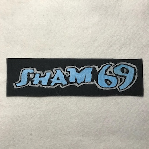 May include: A rectangular black fabric patch featuring the text SHAM 69 in a stylized, light blue font with a thin silver outline. The lettering has a jagged, distressed appearance, and the patch has a woven texture throughout the black background.