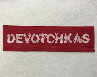 Puede incluir: Un parche rectangular de tela roja con la palabra DEVOTCHKAS impresa en una fuente blanca desgastada. El parche tiene una textura tejida y bordes rectos y limpios, presentando un fuerte contraste entre el fondo rojo intenso y el texto blanco.