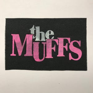 May include: A rectangular black fabric patch featuring the word the in small silver lettering above the word MUFFS in large, bold, pink gradient lettering. The patch has a visible woven texture throughout the black background.