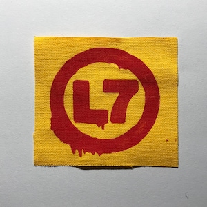 May include: A square fabric patch with a yellow background and a red circular graphic. Inside the red circle, the text L7 is printed in a bold red font. The red circle features a dripping paint effect along the bottom left edge of the design.