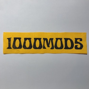 May include: A rectangular fabric patch in a bright yellow color featuring the text 1000MODS printed in a bold, black, stylized font across the center. The patch has a visible woven texture and straight, clean edges on all four sides.