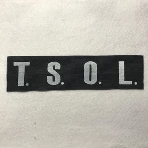 May include: A rectangular black fabric patch featuring the white embroidered text T. S. O. L. in a bold, sans-serif font. The letters are evenly spaced across the horizontal strip, which has a visible woven texture and clean, straight edges.