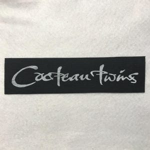 May include: A rectangular black fabric patch featuring the text Cocteau Twins embroidered in a light gray, stylized cursive font. The patch has a woven texture and clean, straight edges, presenting the band name in a clear, legible design.