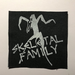 May include: A square black fabric patch featuring a screen-printed graphic in light gray. The design depicts a stylized, angular figure with raised arms, positioned above the text "SKELETAL FAMILY" written in a jagged, distressed font style.
