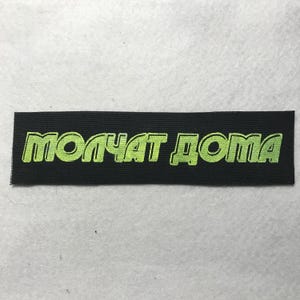 May include: A rectangular black fabric patch featuring the text MOLCHAT DOMA embroidered in a bright lime green thread. The lettering is stylized with a bold, blocky font and a distinct outline, creating a high-contrast visual design.