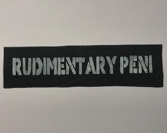 Puede incluir: Un parche rectangular de tela negra con el texto RUDIMENTARY PENI impreso en una fuente blanca de estilo plantilla desgastada en el centro. El parche tiene una textura tejida y bordes rectos y limpios en los cuatro lados.