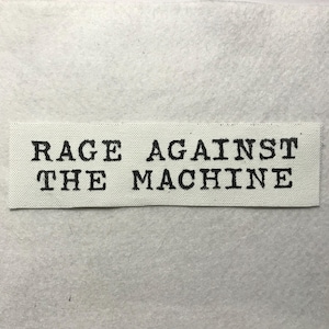 May include: A rectangular off-white fabric patch featuring the black text RAGE AGAINST THE MACHINE printed in a bold, typewriter-style font. The patch has a woven texture and clean, straight edges, presenting a clear and legible design.