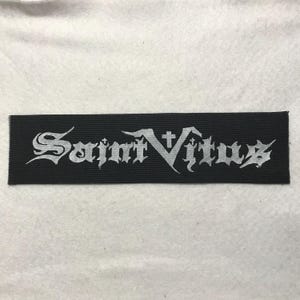 May include: A rectangular black fabric patch featuring the text Saint Vitus embroidered in a white gothic-style font. The lettering includes small cross symbols integrated into the design of the letters, creating a distinct visual graphic.