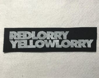 Puede incluir: Un parche rectangular de tela negra con el texto RED LORRY YELLOW LORRY bordado en una fuente gris claro sin remates. El parche tiene una textura tejida y bordes rectos y limpios, mostrando las palabras en dos líneas horizontales.