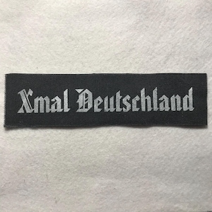 May include: A rectangular black fabric patch featuring the text Xmal Deutschland embroidered in a white gothic-style font. The patch has a woven texture and clean, straight edges, presenting a high-contrast design with clear, legible lettering.