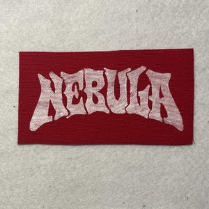 May include: A rectangular fabric patch in a deep red color featuring the word NEBULA printed in a distressed white font across the center. The patch has a woven texture and clean, straight edges, presenting a bold contrast between the red background and the white lettering.