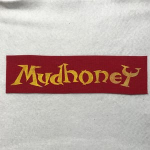 May include: A rectangular fabric patch featuring a deep red background with the word Mudhoney embroidered in a stylized, sharp-edged yellow font. The embroidery has a visible woven texture, and the lettering is centered across the patch.