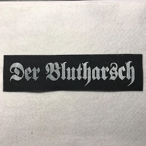 Può includere: Una toppa rettangolare in tessuto nero con il testo bianco Der Blutharsch in un carattere in stile gotico. La toppa presenta una trama tessuta e bordi netti e dritti, offrendo un design ad alto contrasto per l'applicazione su articoli tessili.