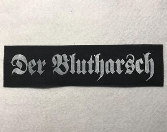 May include: A rectangular black fabric patch featuring the white text Der Blutharsch in a gothic-style font. The patch has a woven texture and clean, straight edges, presenting a high-contrast design suitable for application onto various textile items.