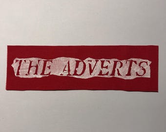 May include: A rectangular red fabric patch featuring the text THE ADVERTS printed in white. The lettering has a distressed, weathered appearance with uneven edges, set against a solid, vibrant red background with a visible woven texture.