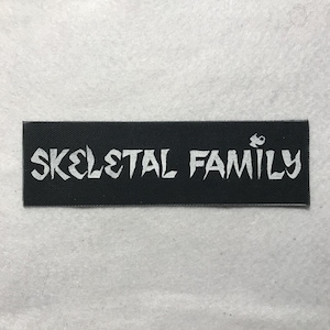 Puede incluir: Un parche rectangular negro con las palabras "SKELETAL FAMILY" en blanco, con letras estilizadas. Un pequeño gráfico blanco está encima de la palabra "FAMILY". El parche es para ropa o accesorios.