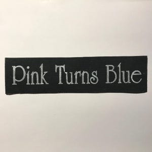 Może przedstawiać: Prostokątna czarna naszywka z tkaniny z napisem Pink Turns Blue nadrukowanym srebrną czcionką szeryfową. Naszywka ma widoczną tkaną fakturę oraz czyste, proste krawędzie, co tworzy projekt o wysokim kontraście.