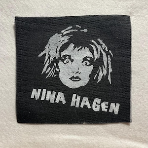 Könnte beinhalten: Schwarzer quadratischer Aufnäher mit einem Graustufenporträt von Nina Hagen. Der Name "NINA HAGEN" ist in weißen, fetten Großbuchstaben darunter gedruckt. Der Aufnäher hat eine gewebte Textur.