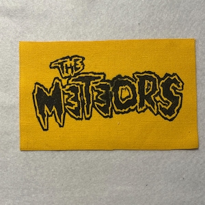 May include: A rectangular yellow fabric patch featuring the text THE METEORS printed in a bold, jagged black font. The design has a textured appearance with visible woven fabric threads throughout the surface of the patch.