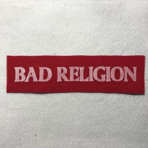 May include: A rectangular red fabric patch featuring the text BAD RELIGION embroidered in a light pink, serif font. The patch has a woven texture and clean, straight edges, presenting a bold, high-contrast design suitable for attachment to clothing.