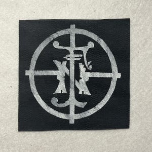 Op de afbeelding: Een vierkante zwarte stoffen patch met een wit bedrukt ontwerp. De afbeelding bestaat uit een grote cirkel die wordt doorkruist door een verticale en een horizontale lijn, waardoor een kruisvorm ontstaat. In het midden staan gestileerde letters.
