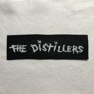 May include: A black rectangular patch featuring the white text "THE DISTILLERS". The text is in a stylized font. The patch is made of woven fabric, possibly a clothing label or patch.