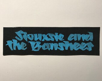 May include: A rectangular black fabric patch featuring the text Siouxsie and the Banshees embroidered in a bright blue, stylised, angular font. The patch has a woven texture and clean, straight edges, displaying the band name clearly.